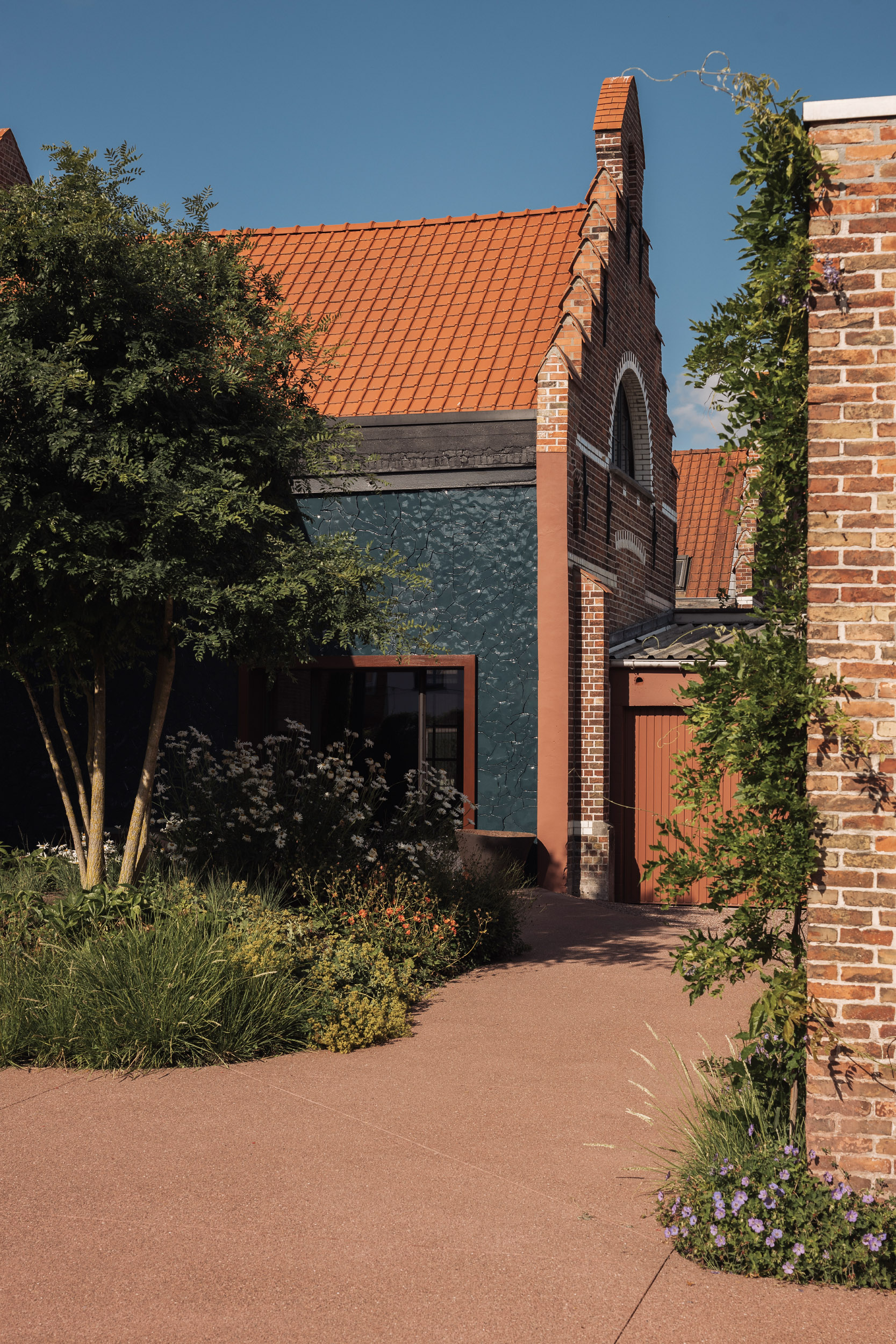 Facade of a rustic house with a textured dark green wall, red-framed window, brick elements, and lush garden in the foreground.