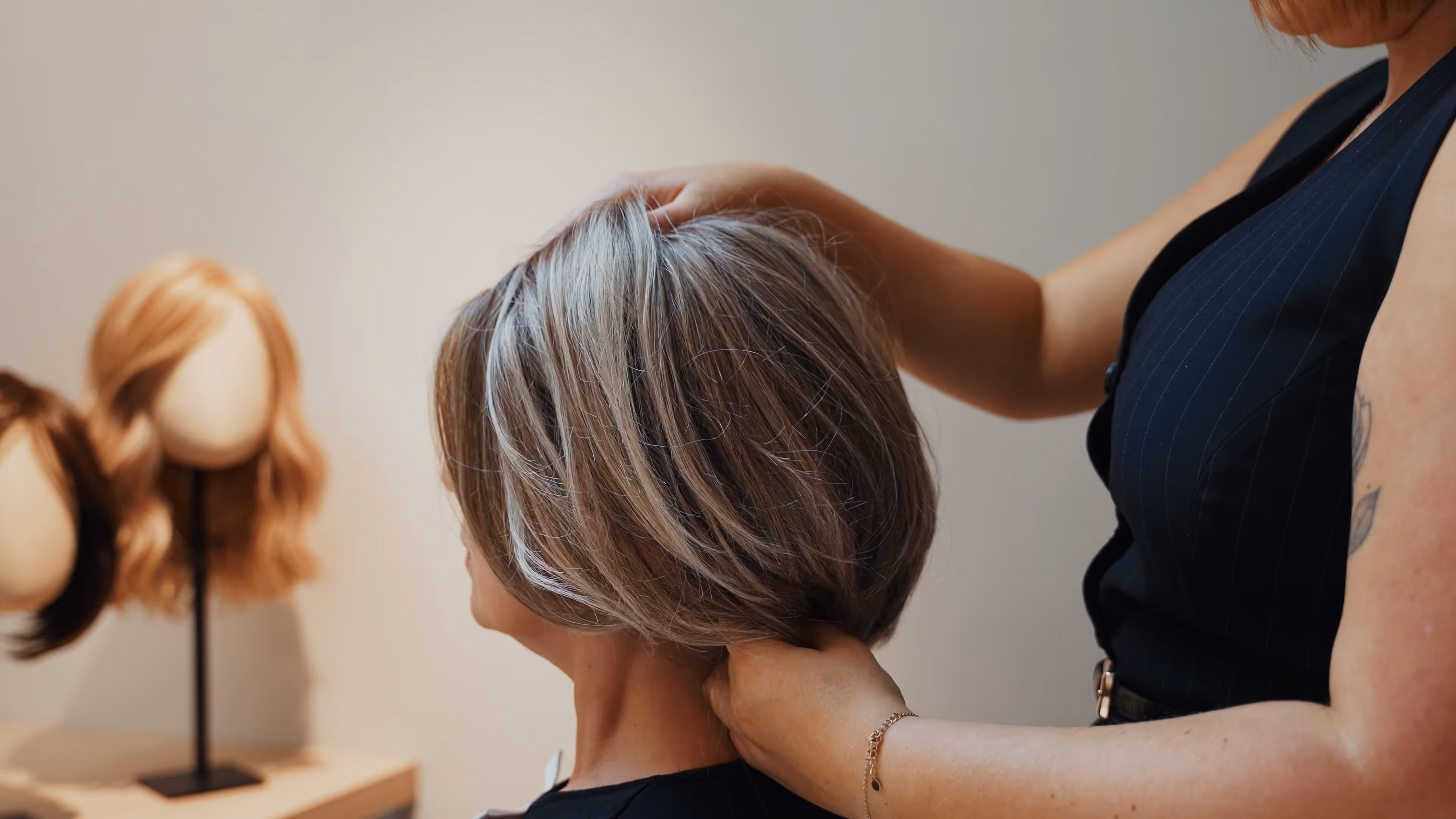 Persoon die een kort grijs bobkapsel stylt bij een zittende vrouw, met pruiken op mannequinhoofden op de achtergrond.