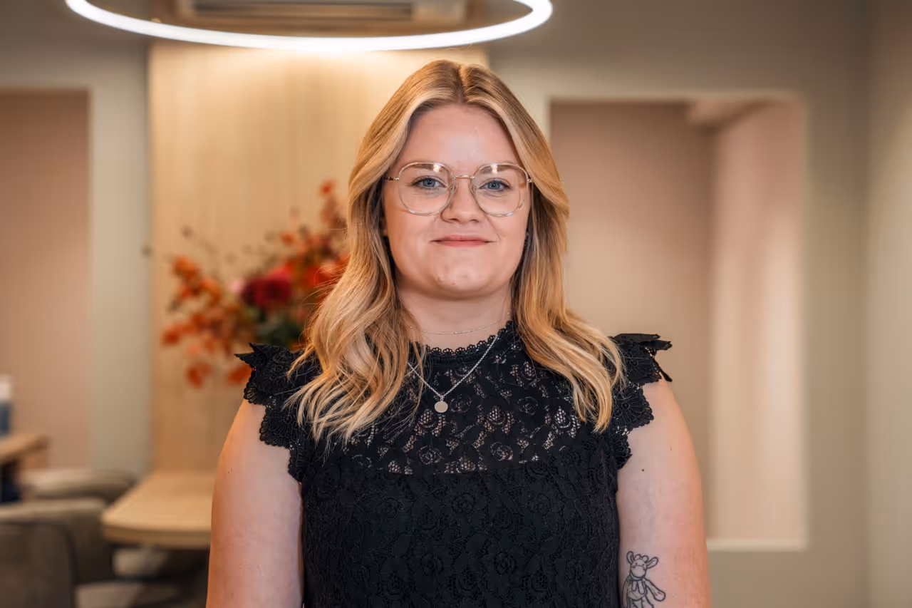 Kapster Anaïs glimlacht naar de camera; draagt zwarte blouse met kant, heeft blond haar.