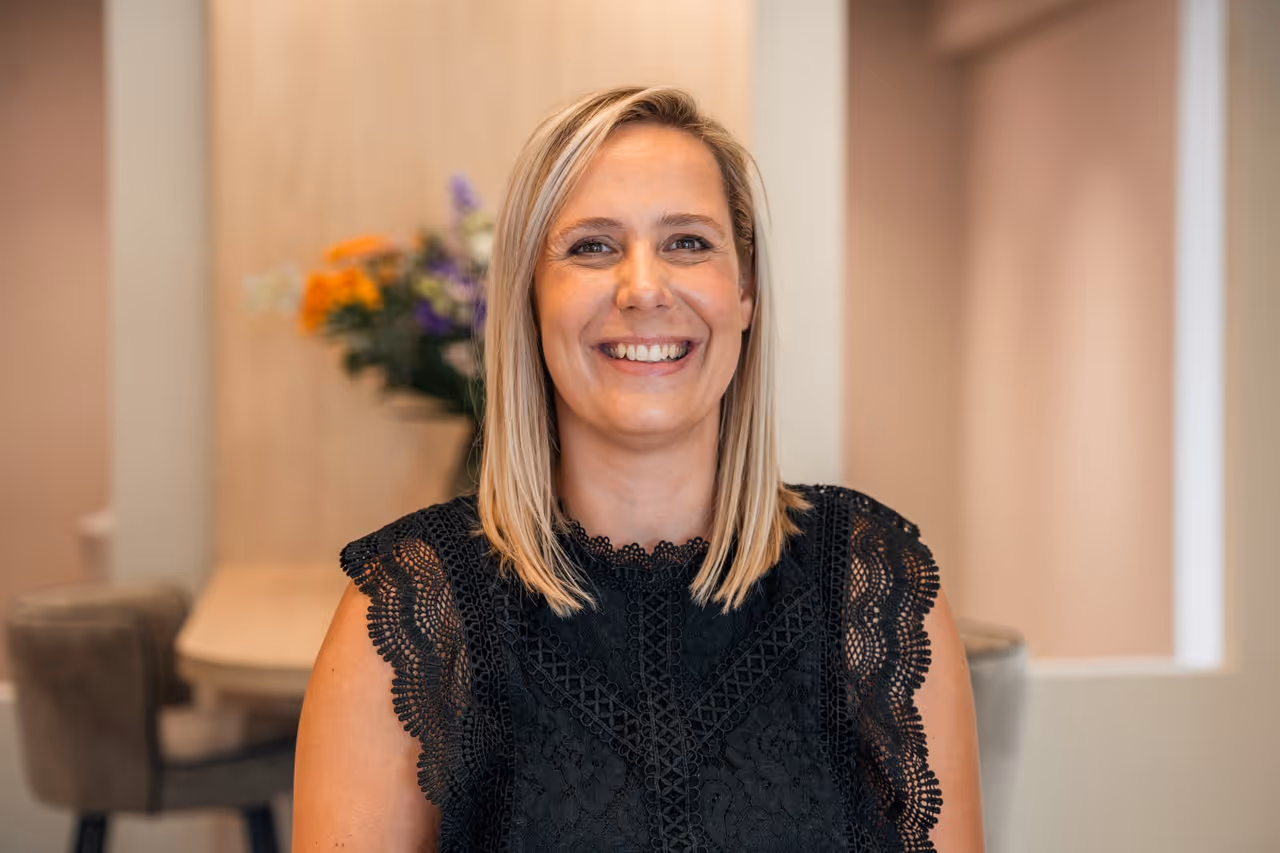 Kapster Miranda glimlacht naar de camera; draagt zwarte blouse, heeft blond haar.