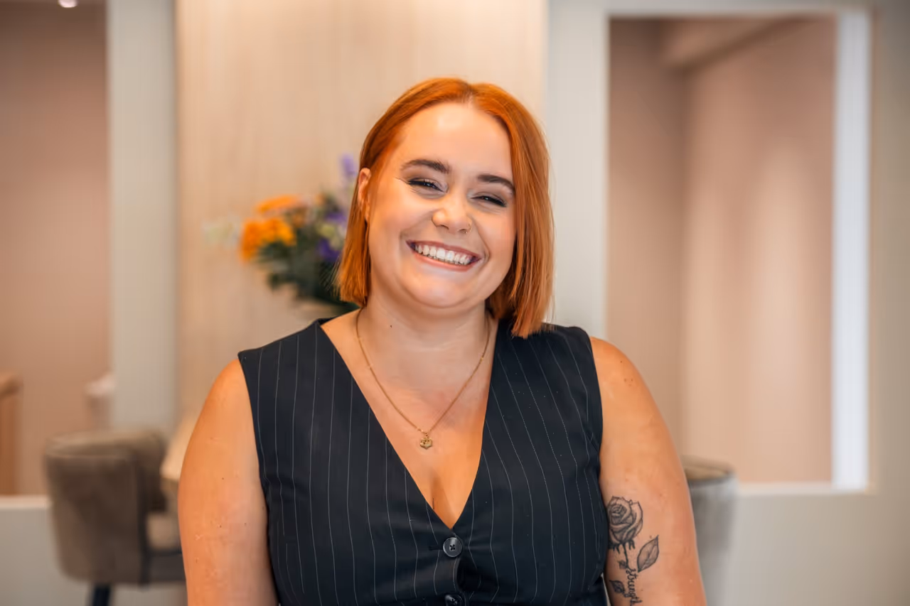 Kapster Nikky glimlacht naar de camera; draagt zwarte blouse met strepen, heeft rood haar.