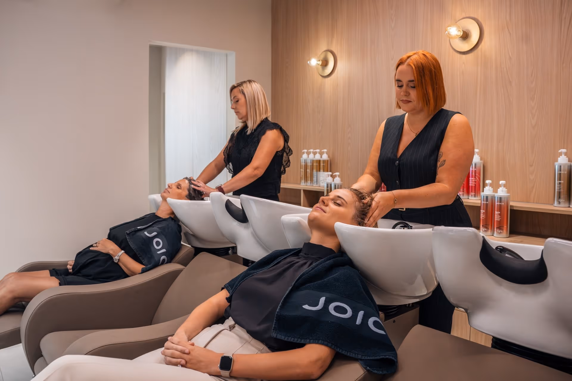 Twee vrouwen laten hun haar wassen door kapsters in een modern salon met houten wanden en planken vol haarproducten.