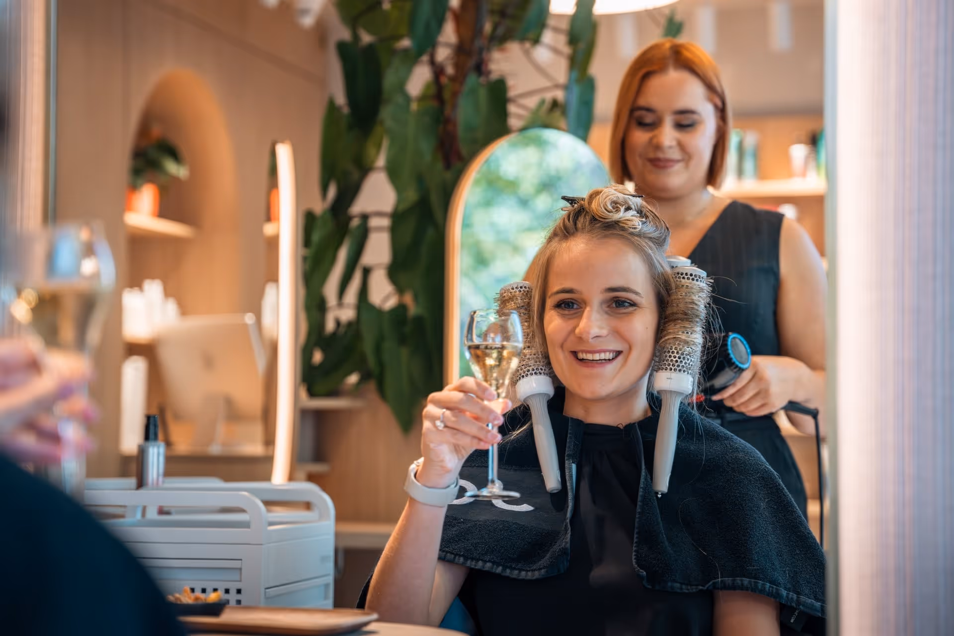 Een glimlachende vrouw heft een glas witte wijn in een kapsalon terwijl een kapper haar haar föhnt.