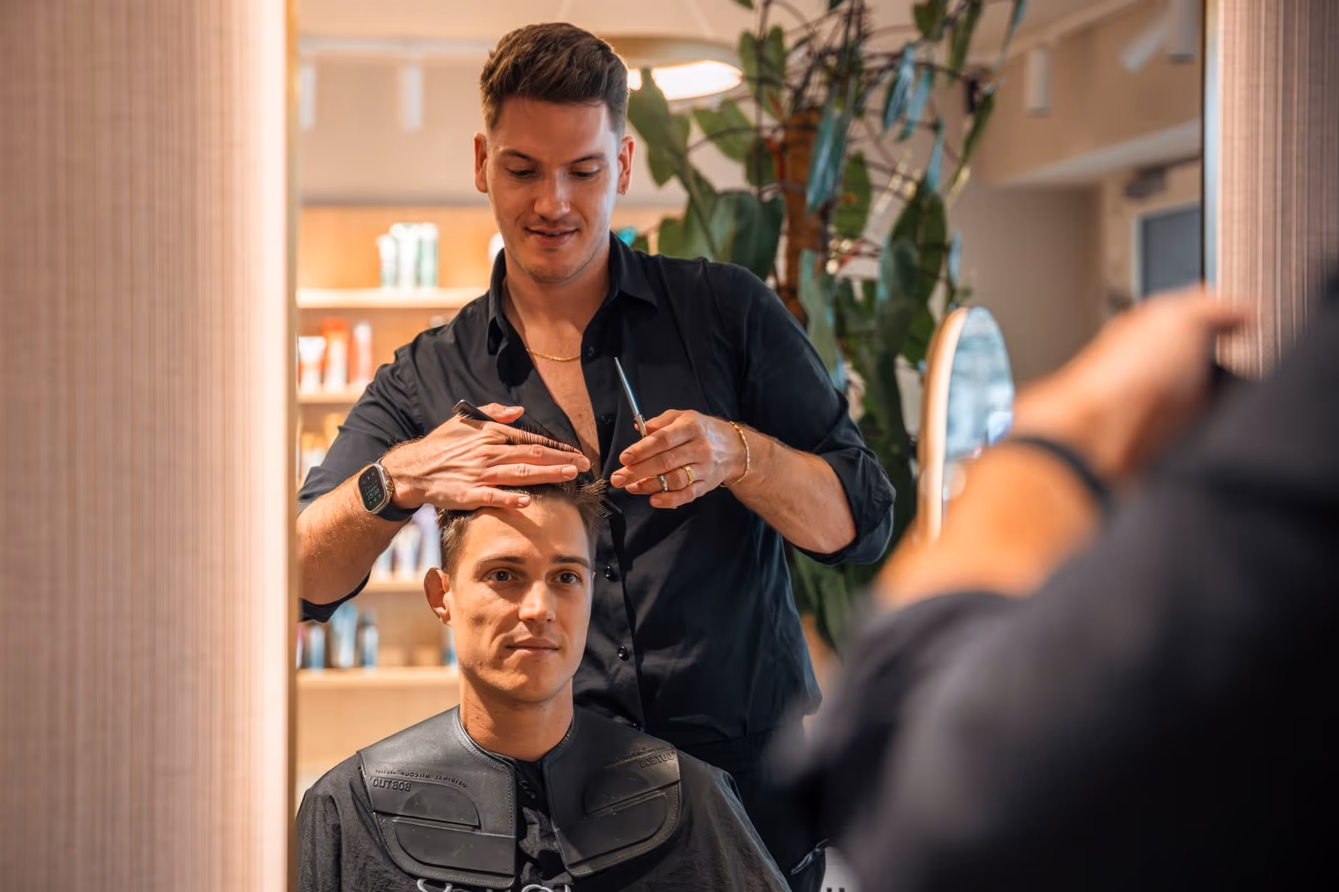 Kapper in zwarte blouse knipt het haar van een man in een kapsalon met planten op de achtergrond.