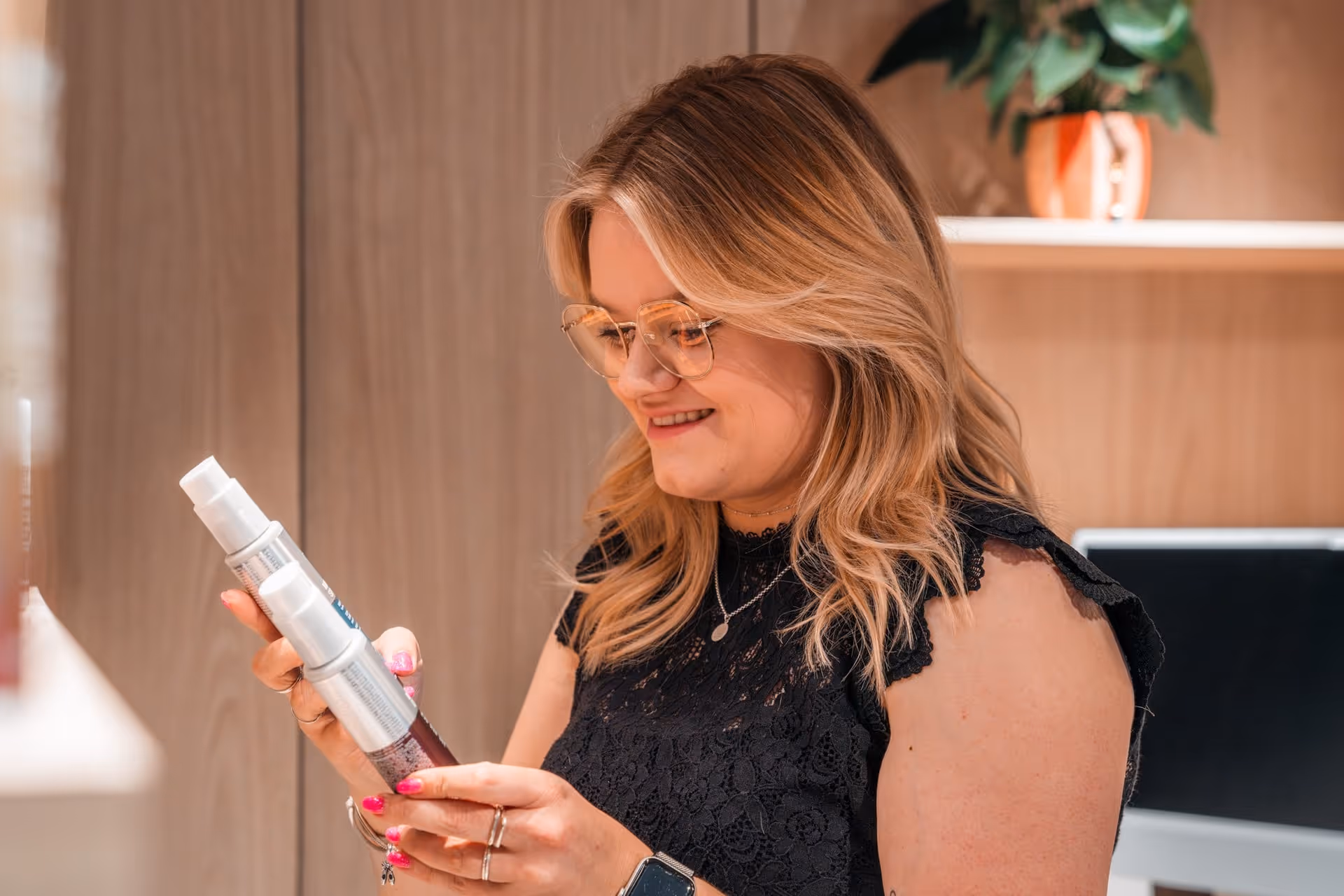 Glimlachende vrouw met blond haar en bril bekijkt twee haarproducten.