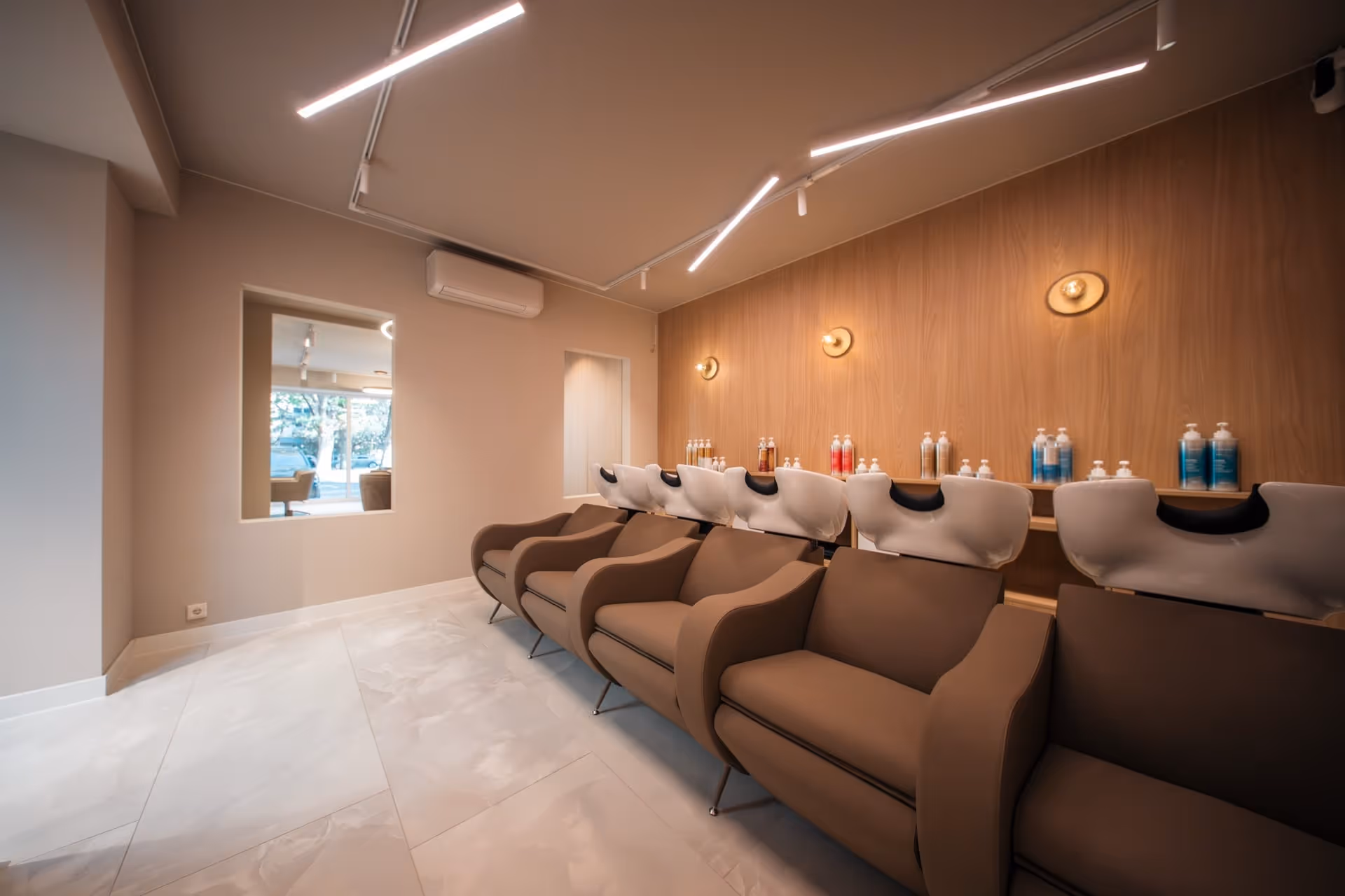 Moderne shampoo-ruimte in een kapsalon met vijf bruine wasstoelen tegen een houten wand, met flessen haarproducten op planken erboven.