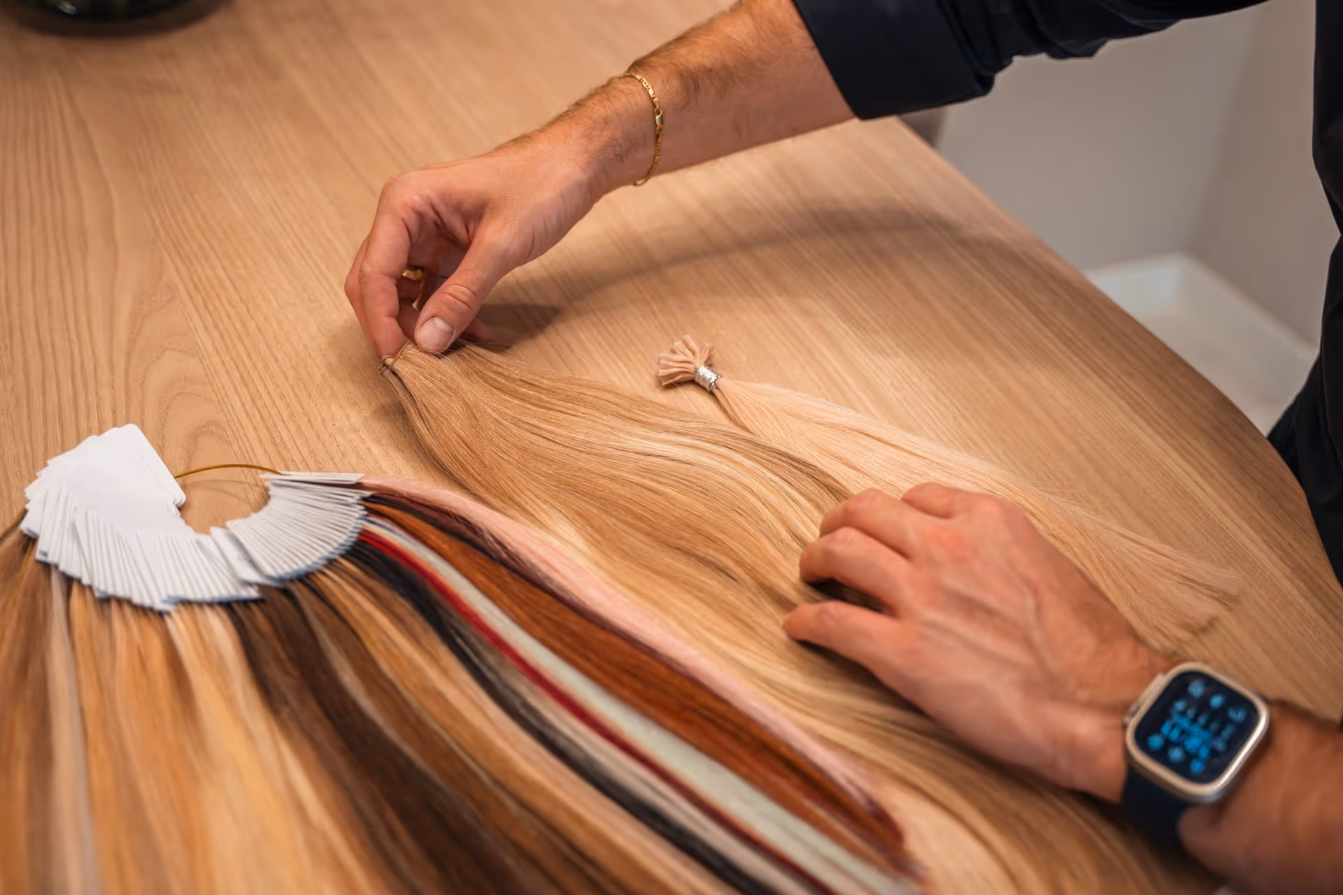 Handen die blonde haarextensies ordenen op een houten tafel naast een kleurenstaal met verschillende haarkleuren.