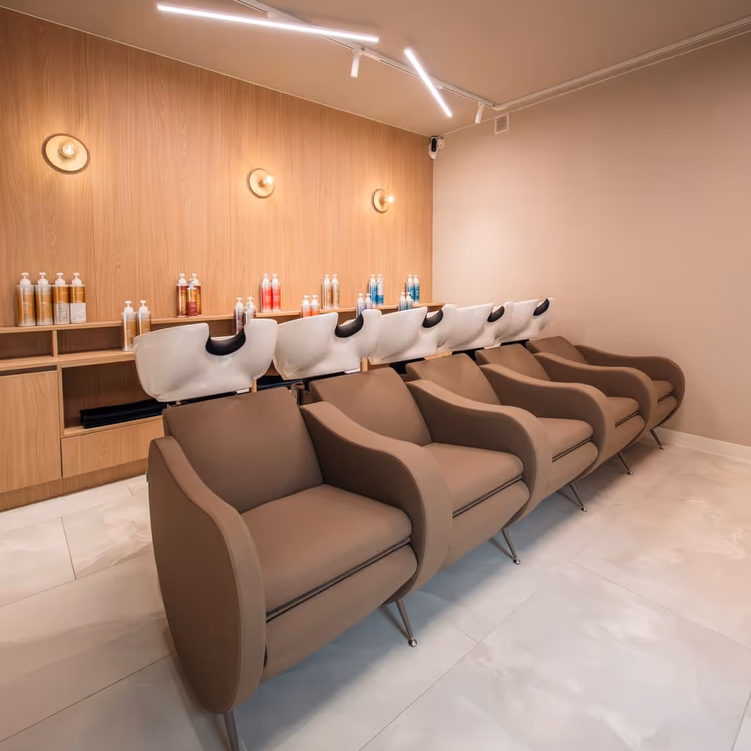 Rij van vijf bruine kappersstoelen met witte wasbakken in een modern kapsaloninterieur met houten lambrisering en wandlampen.