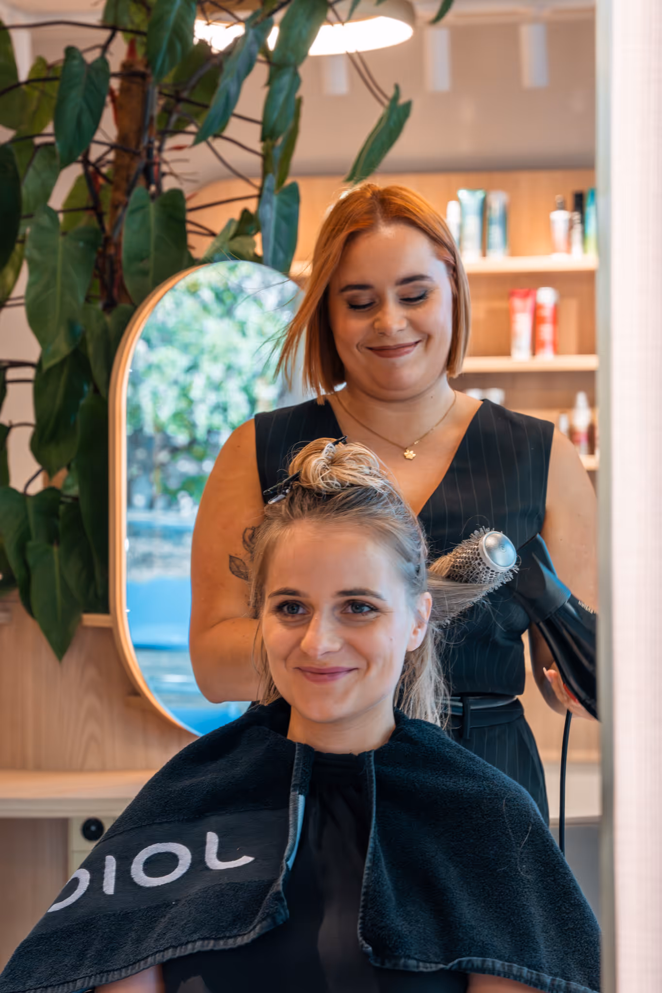 Kapper die het haar van een glimlachende vrouw droogt met een ronde borstel en een föhn in een kapsalon.