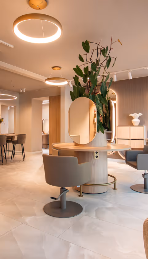 Modern saloninterieur met beige stoelen rond een ronde tafel, grote groene plant, ronde spiegel en warme ringvormige plafondlampen.