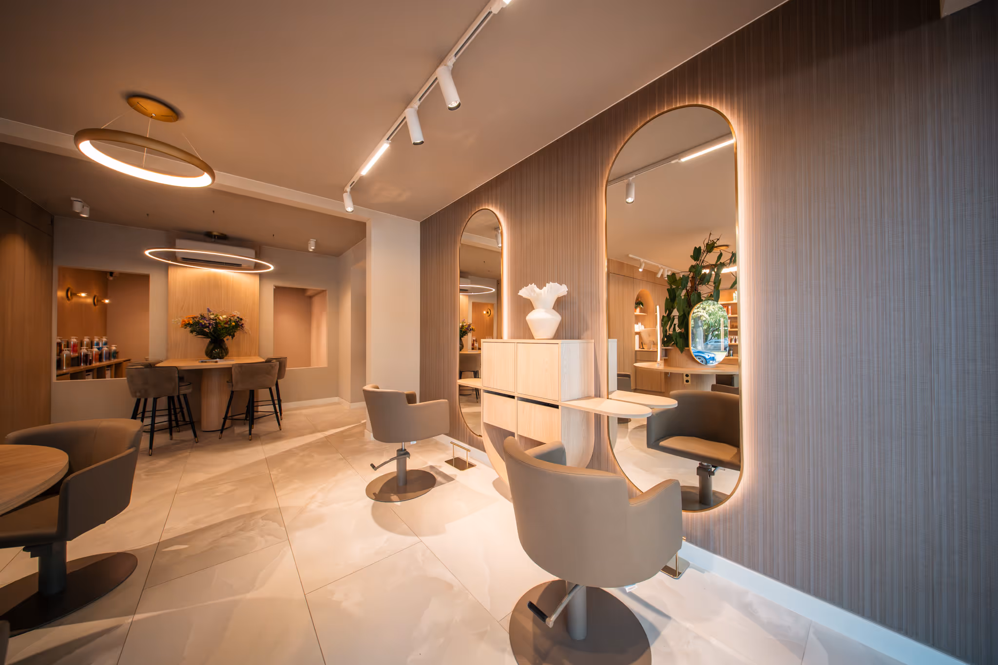 Modern saloninterieur met beige kappersstoelen voor grote ovale spiegels en een tafel met een vaas met bloemen op de achtergrond.