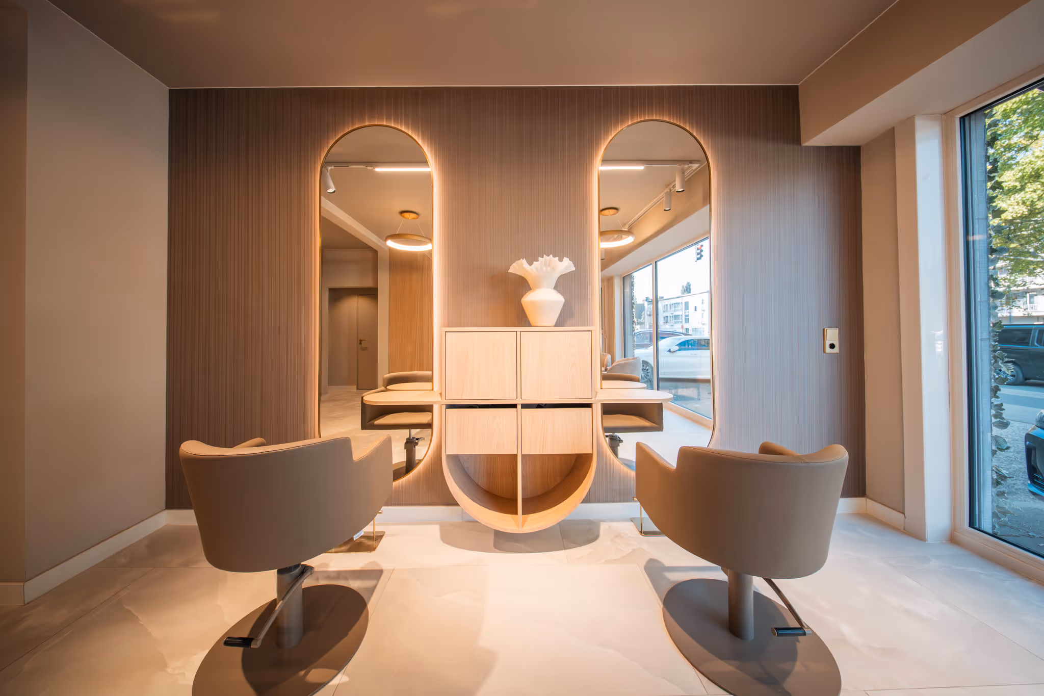 Modern saloninterieur met twee beige kappersstoelen tegenover ovale spiegels met achtergrondverlichting en een houten kast met een witte decoratieve vaas.