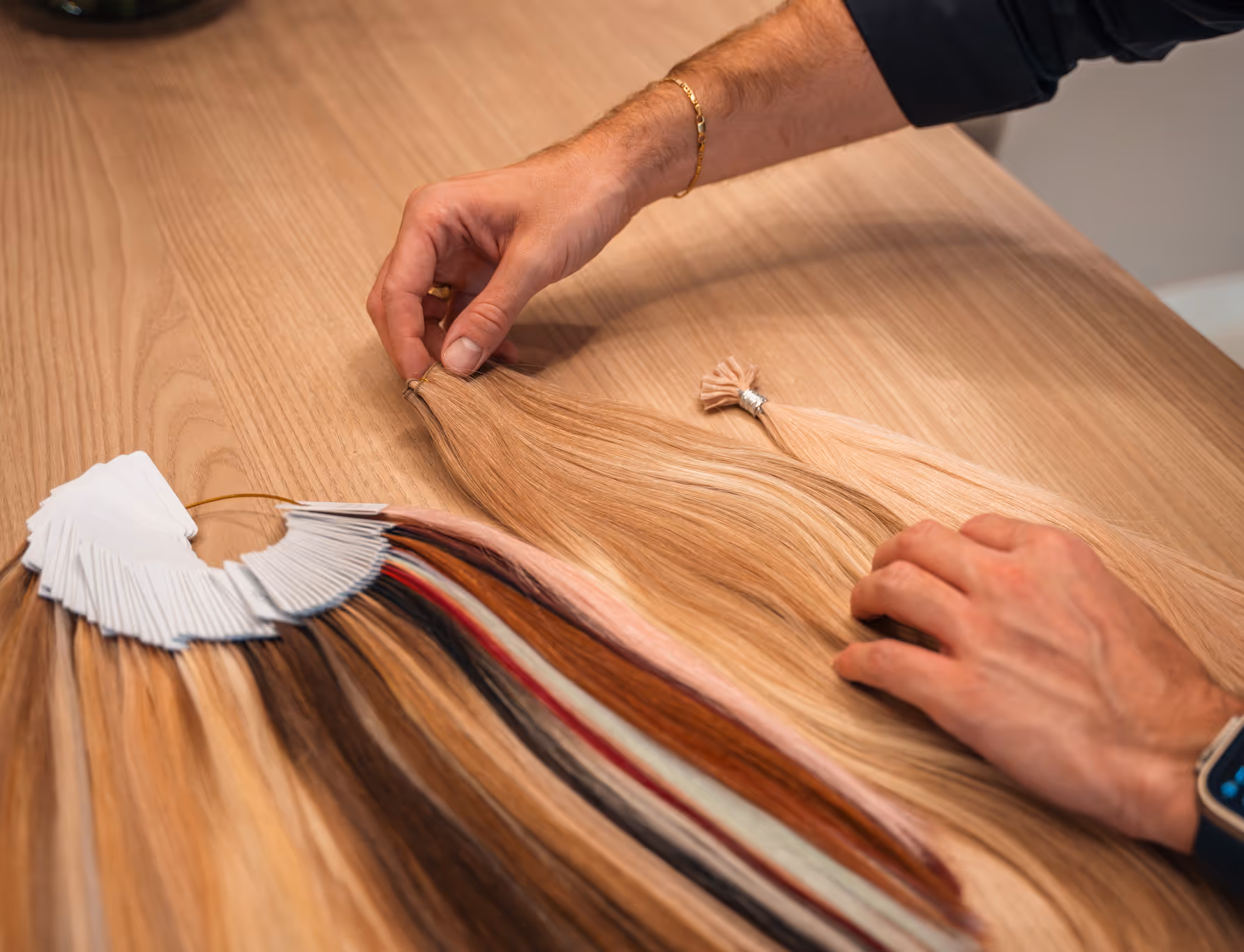 Hands selecting a light brown hair extension from a variety of hair color samples on a wooden table.