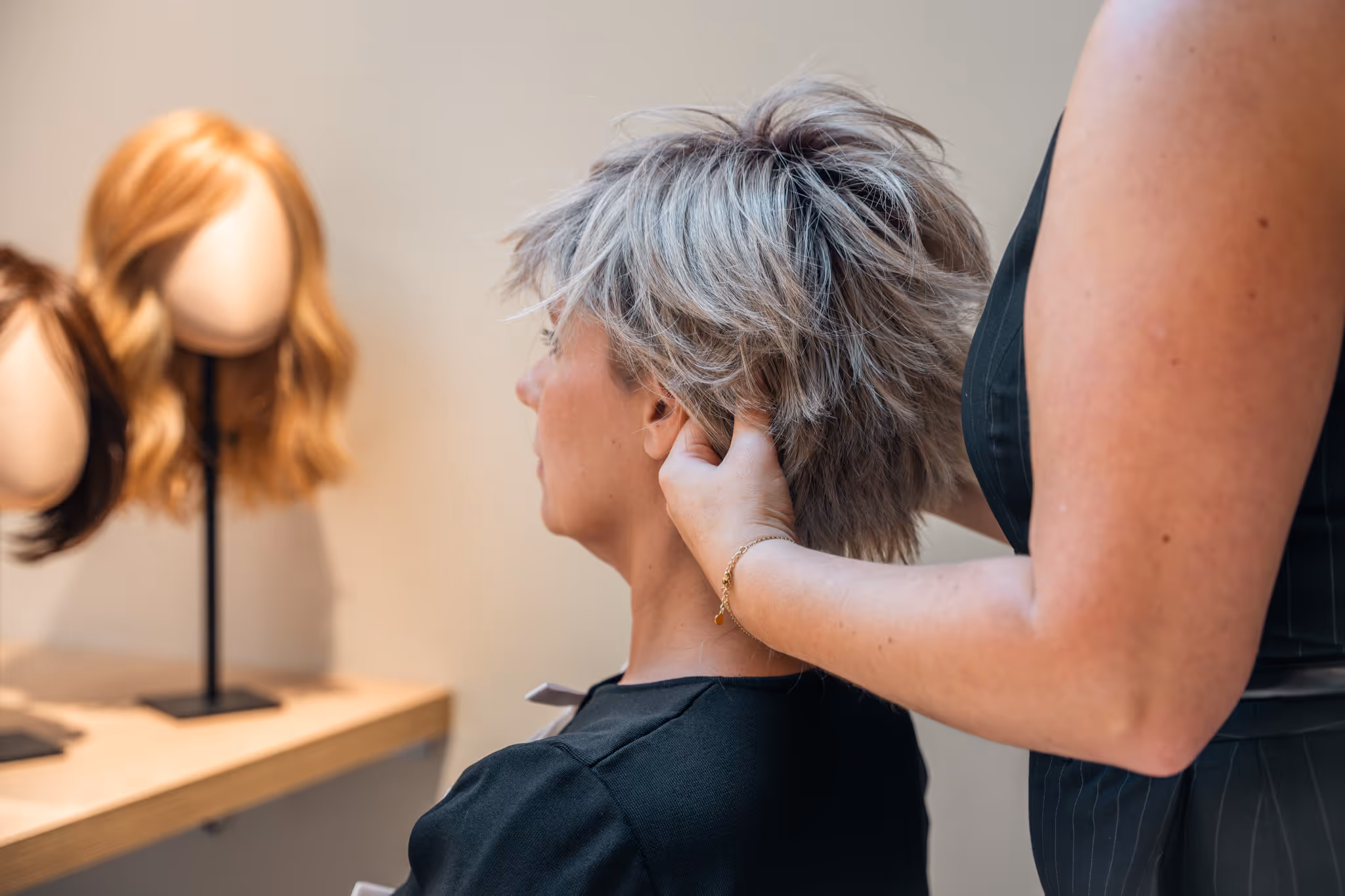 Kapper stylt een kort gelaagd grijs haarwerk van een zittende vrouw met mannequinhoofden met pruiken op de achtergrond.