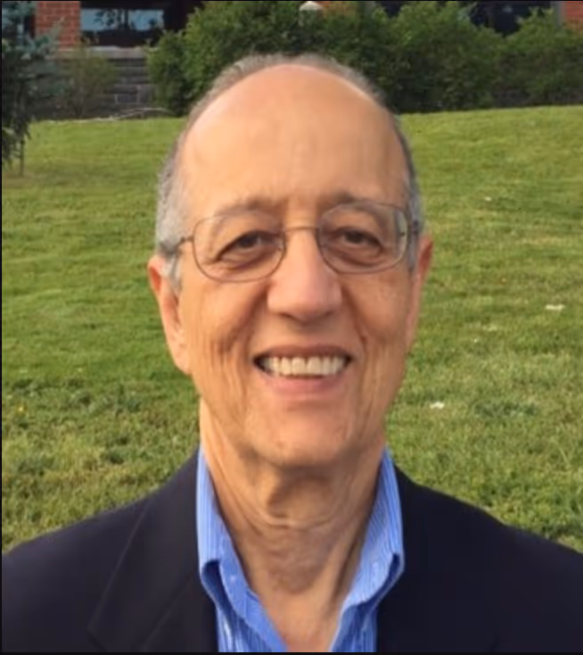 A smiling man outdoors in a grassy area. He is wearing glasses, a blue collared shirt, and a dark blazer. The man has short, greying hair, and his teeth are visible as he smiles warmly. The background includes a well-maintained lawn, with a few scattered leaves and plants, and there is a hint of a brick building and trees in the distance.