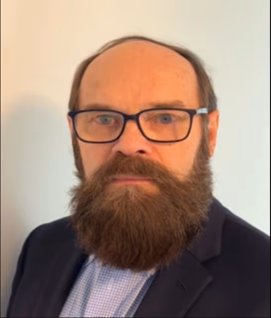 A professional head-and-shoulders portrait of a man against a plain, light-colored background. He has fair skin, a receding hairline, and short brown hair on the sides. He is wearing rectangular, blue-rimmed glasses and has a full, thick brown beard that extends several inches below his chin, covering his entire neck area. He is dressed in a dark suit jacket over a light blue checkered shirt.