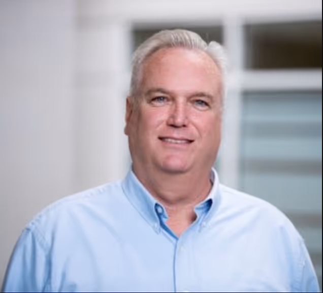 A middle-aged man with short, light gray hair and a light complexion. He is facing the camera and smiling slightly. He is wearing a light blue button-down shirt with a collar. The background is softly focused, consisting of a light-colored wall and a window with horizontal blinds, suggesting an indoor setting, perhaps an office or a modern, professional environment.