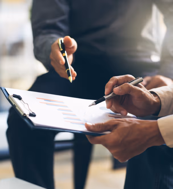 A close-up of two individuals who are engaged in a discussion and examining a document on a clipboard. Both individuals are seated, with the focus on their hands and the document rather than their faces or clothing. The person on the left, dressed in a dark shirt, holds a pen and points towards the document, while the person on the right, dressed in a light-colored shirt, holds another pen. The document on the clipboard displays various colorful bar graphs and possibly some textual data, though the text is not clearly visible. The background is softly blurred, indicating an indoor environment with natural light filtering in.