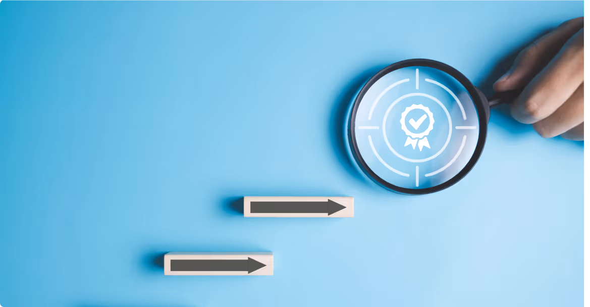 Light blue background with a magnifying glass held by a hand on the right side. The lens of the magnifying glass focuses on a white target symbol featuring a circular badge with a checkmark in the center. To the left of the magnifying glass are two wooden blocks, each with an arrow pointing to the right. The arrows on the blocks are gray, and the blocks are positioned one above the other, slightly staggered.