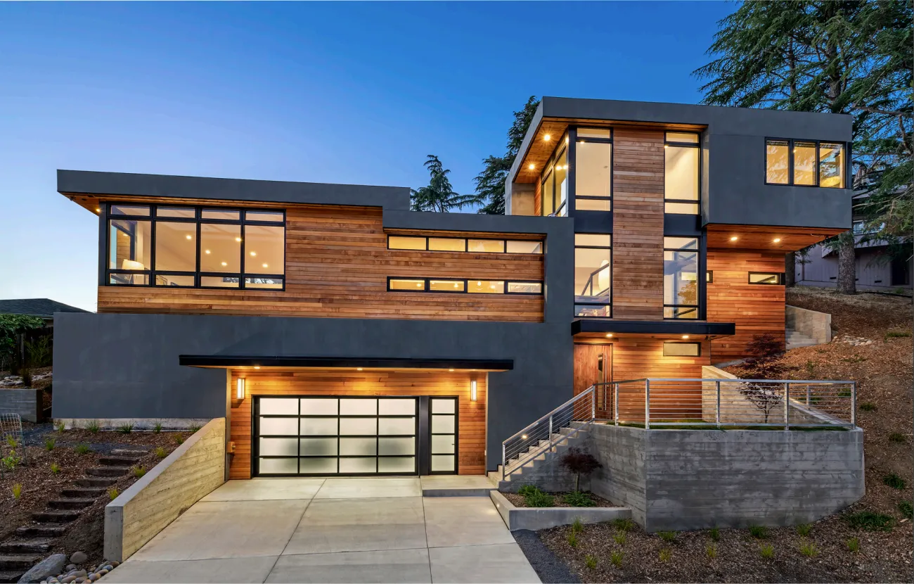 Modern two-story house with wooden and gray exterior, large windows, and a lit garage at dusk.