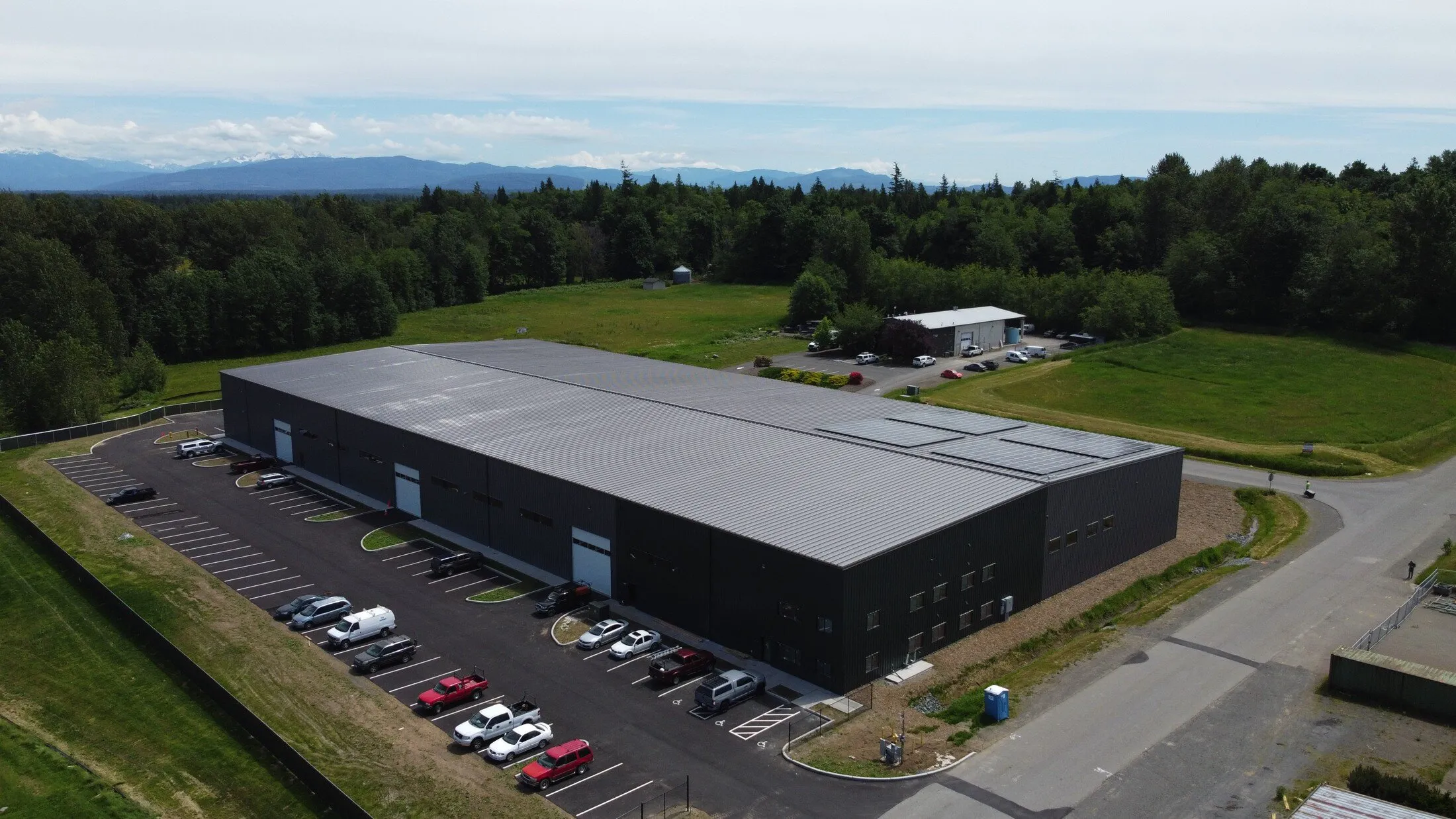 A large industrial warehouse with a corrugated metal roof and multiple parking spaces, surrounded by greenery and distant mountains under a partly cloudy sky.