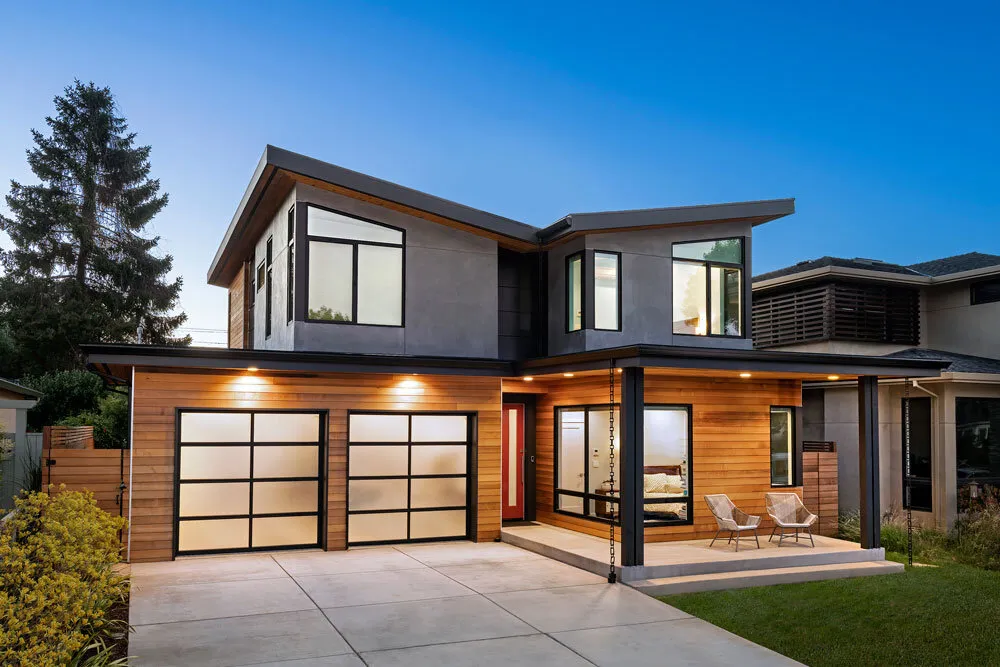 Modern two-story house with wood siding, large windows, and a lit front porch with two chairs.