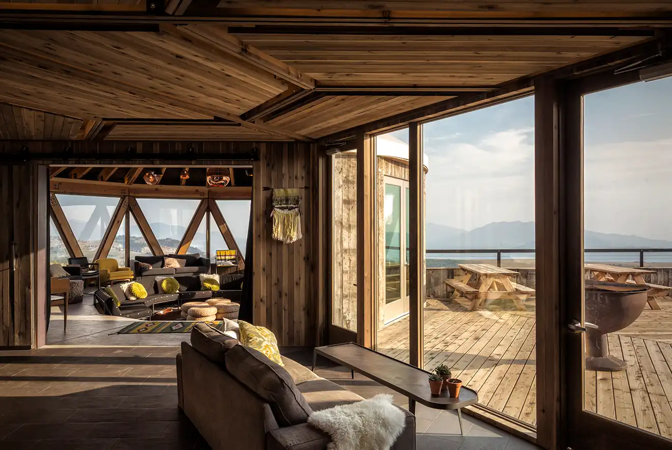 Interior view of a cozy wooden lodge with a sofa, large windows overlooking a deck with picnic tables and mountains in the distance.