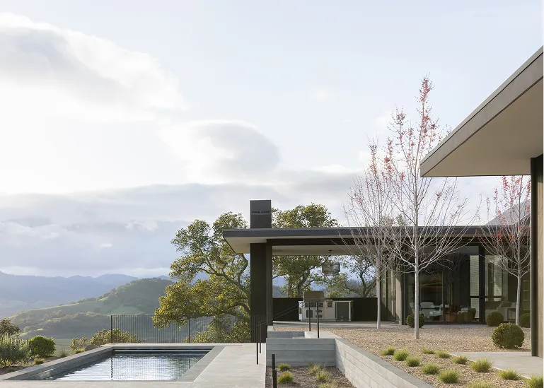 Modern house with large glass windows, a patio, and an infinity pool overlooking a landscape of green hills under a cloudy sky.