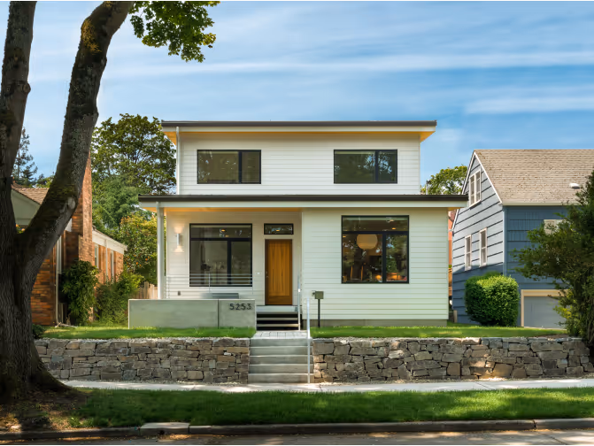 Modern two-story white house with large black-framed windows, wooden front door, and stone retaining wall.