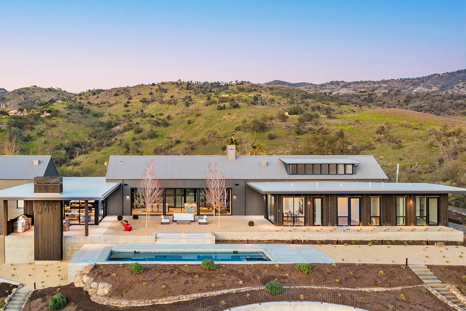 Modern single-story house with large glass doors, outdoor seating area, and a narrow swimming pool, set against rolling green hills at sunset.