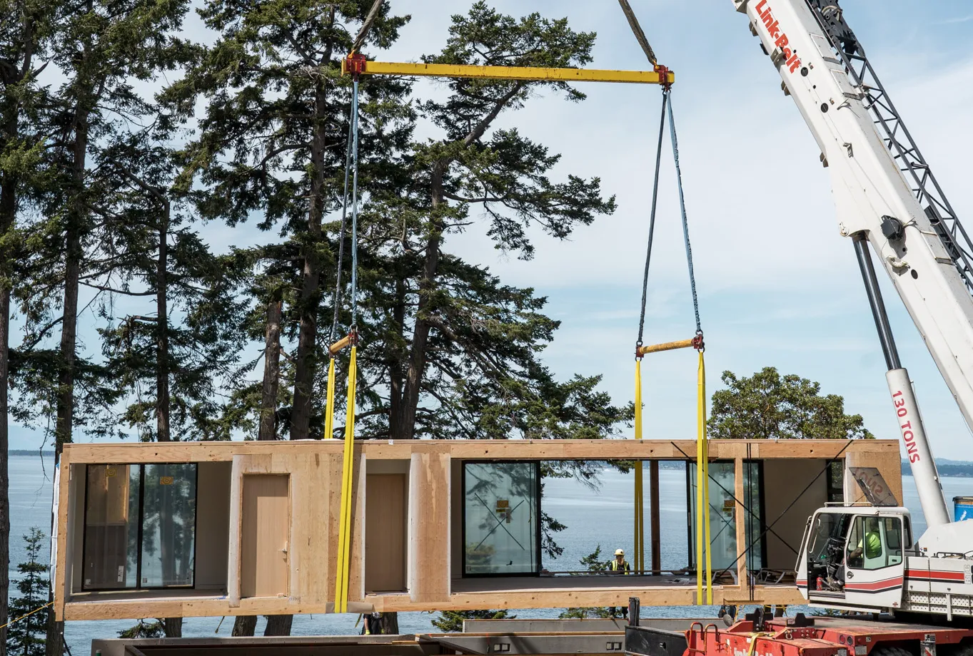 A crane lifts a large prefabricated modular building section near a forested waterfront.