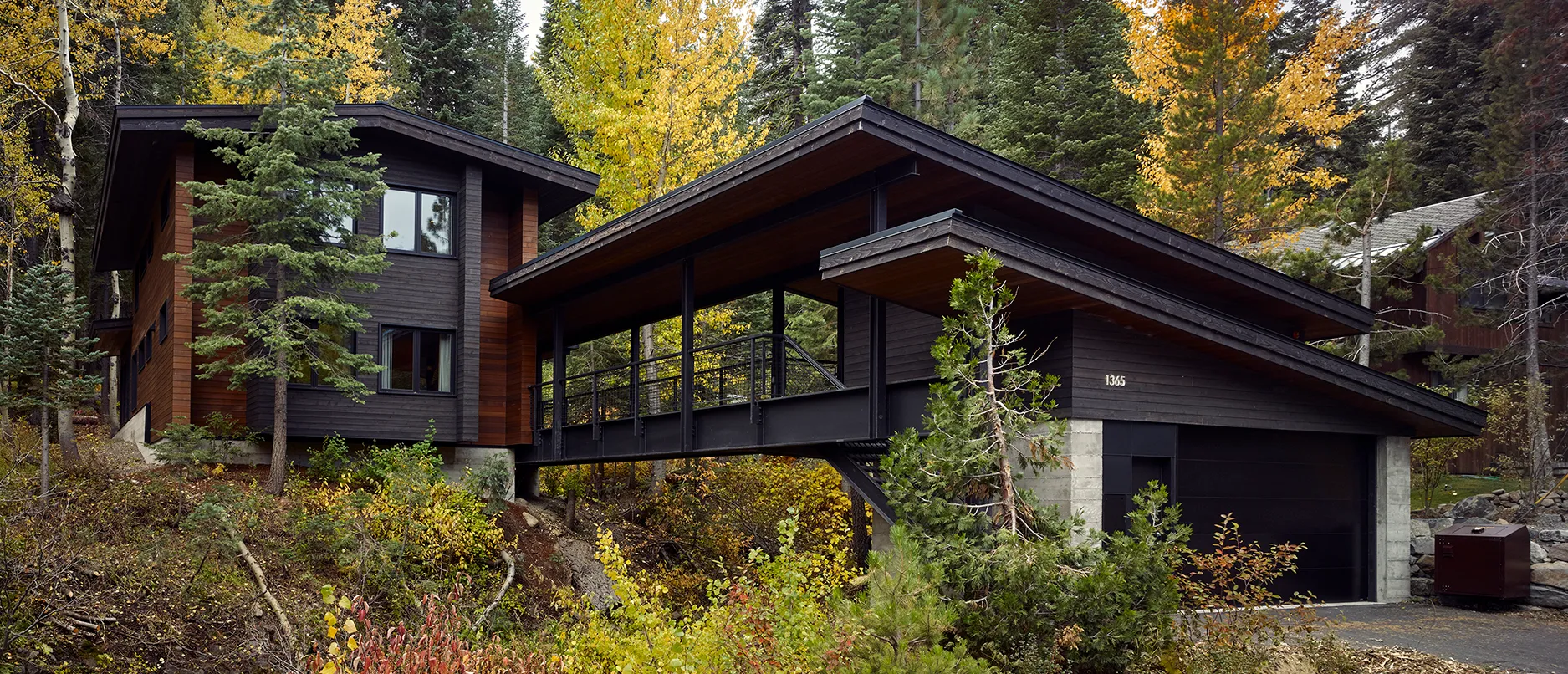 Modern black and wood house surrounded by autumn trees with a covered bridge connecting two parts over a sloped landscape.