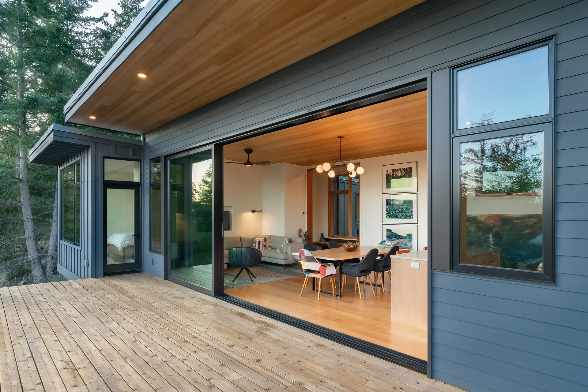 Modern house exterior with large sliding glass doors opening to a wooden deck, showing a dining area and living room inside.