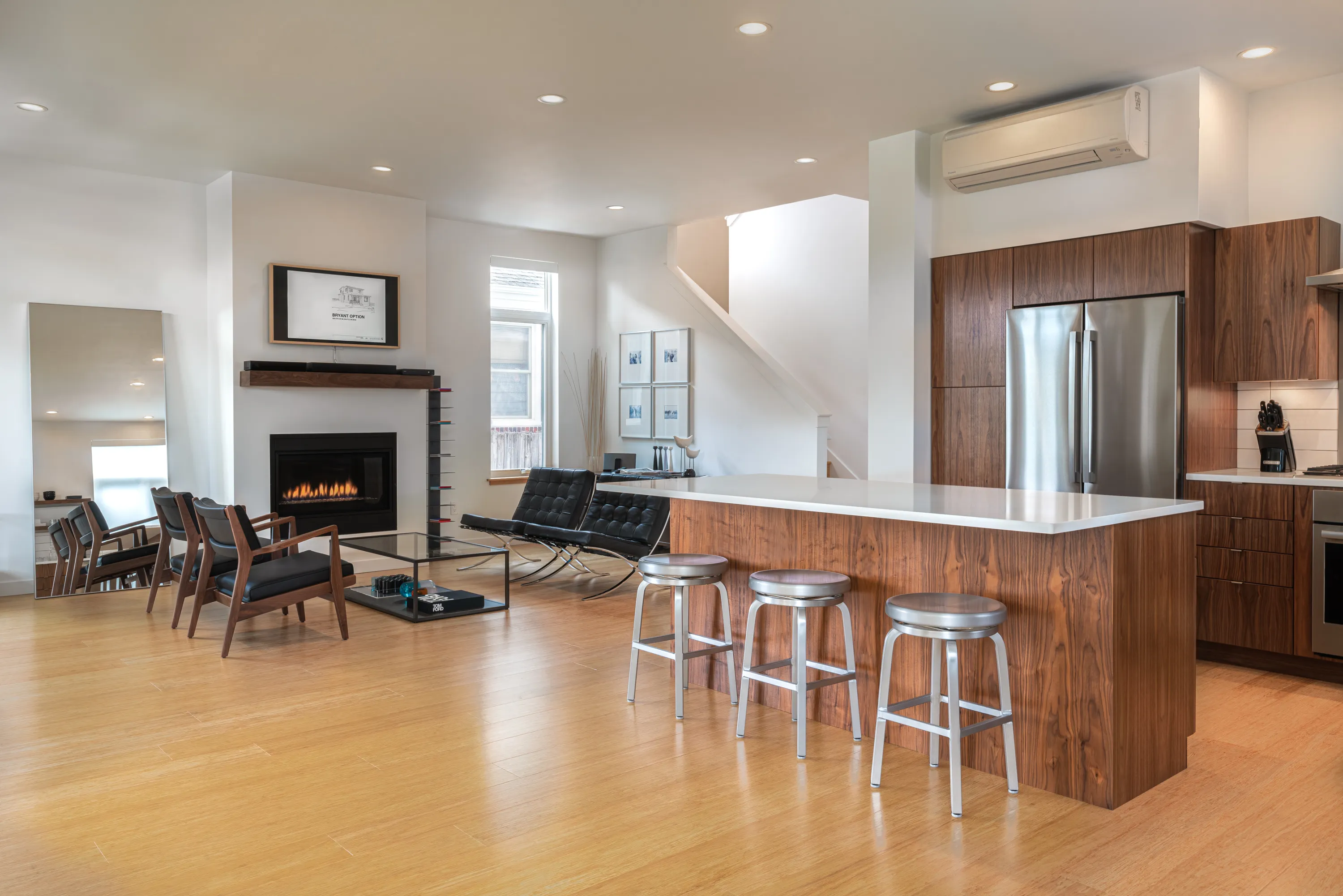 Modern open living room and kitchen with wood flooring, a fireplace, black chairs, a wooden kitchen island with three metal stools, and stainless steel appliances.