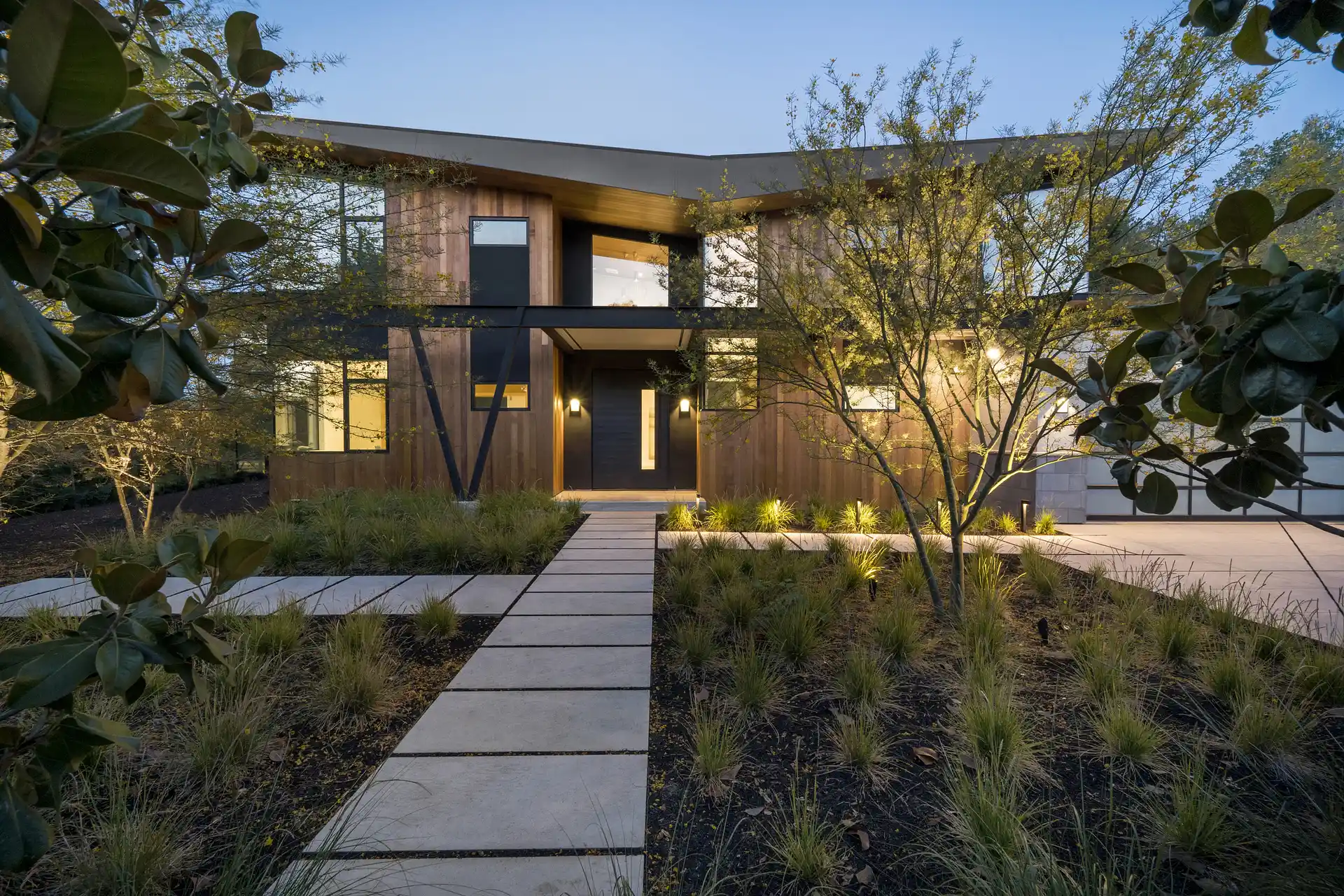 Modern two-story house with wood siding, large windows, and illuminated landscaping at dusk.