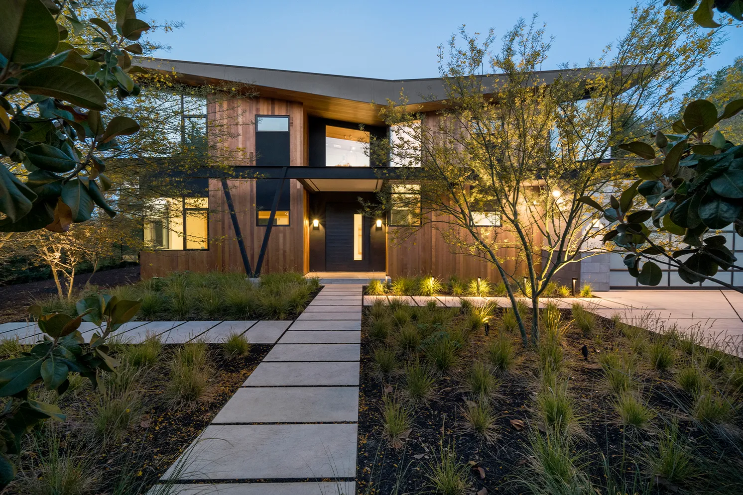 Modern two-story house with wood siding, large windows, and illuminated landscaping at dusk.