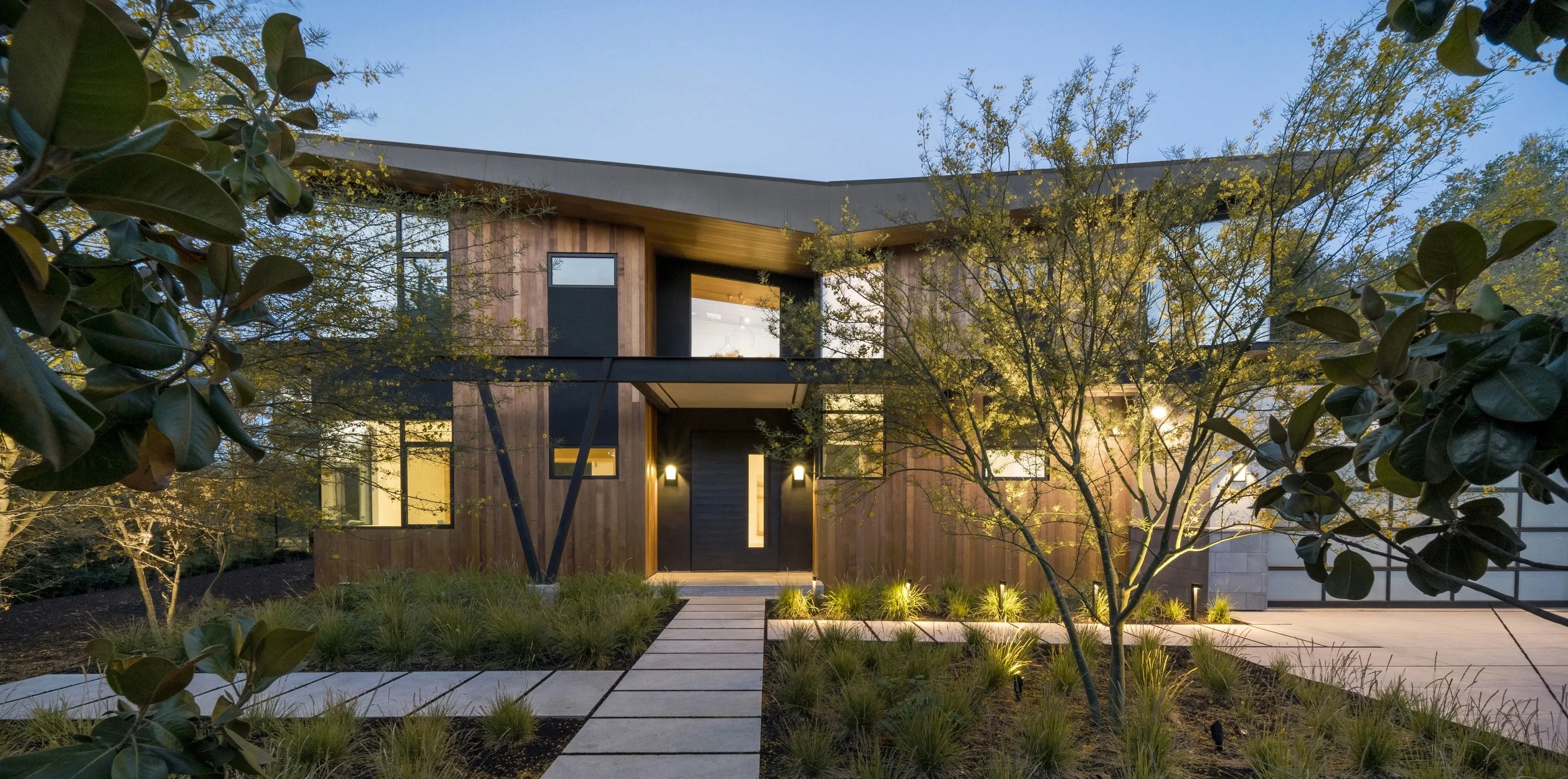 Modern two-story house with wood siding, large windows, and illuminated landscaping at dusk.