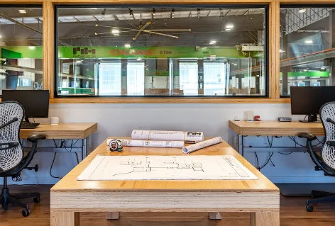 Wooden table with architectural blueprints and rolled plans in a modern office with two desks and office chairs beside large window overlooking factory.