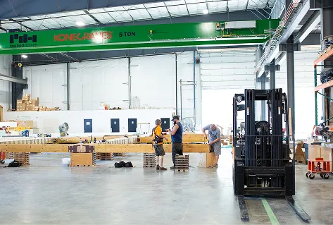 Three workers handling a long wooden beam inside a spacious industrial warehouse with a green overhead crane.