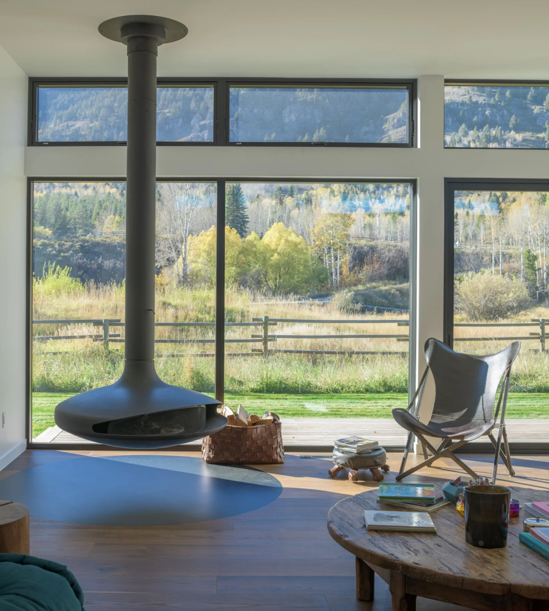 Modern living room with suspended black fireplace, woven basket of firewood, butterfly chair, rustic wooden coffee table, and large windows showing a fenced yard and autumn trees.
