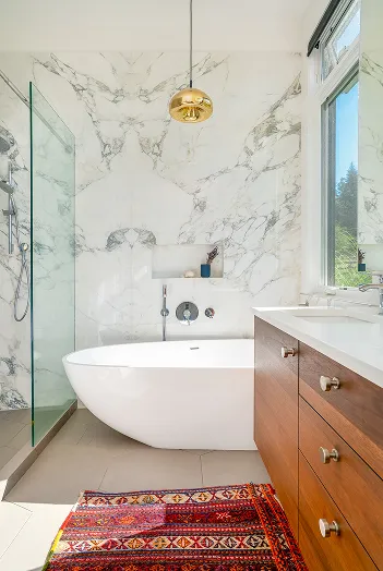 Modern bathroom with a white freestanding bathtub against marble walls, glass shower enclosure, wooden vanity with drawers, and a colorful patterned rug on the floor.