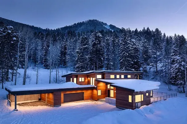 Modern wooden house with snow-covered roof and warm interior lights, surrounded by snow and tall pine trees at dusk.