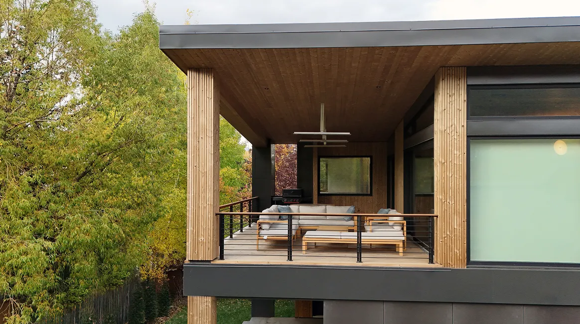Modern covered balcony with wooden pillars, gray cushioned seating, and a coffee table surrounded by green trees.