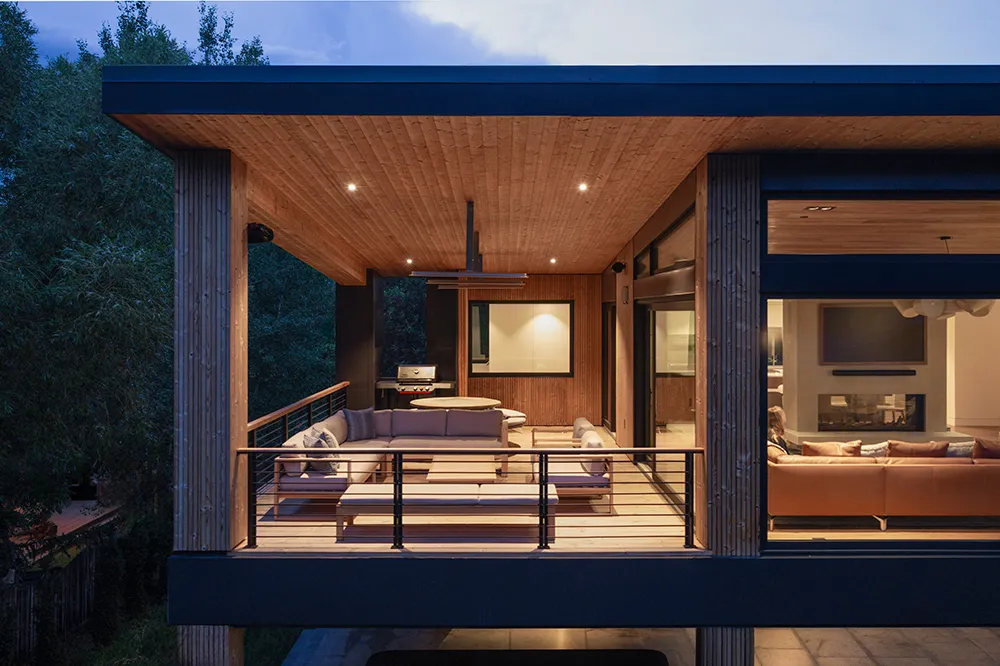 Modern covered balcony with wooden pillars, gray cushioned seating, and a coffee table surrounded by green trees.