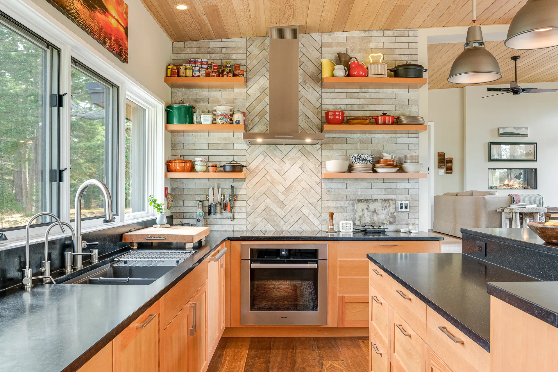 Modern kitchen with wooden cabinets, black countertops, large windows, open shelves with kitchenware, and stainless steel oven and sink.