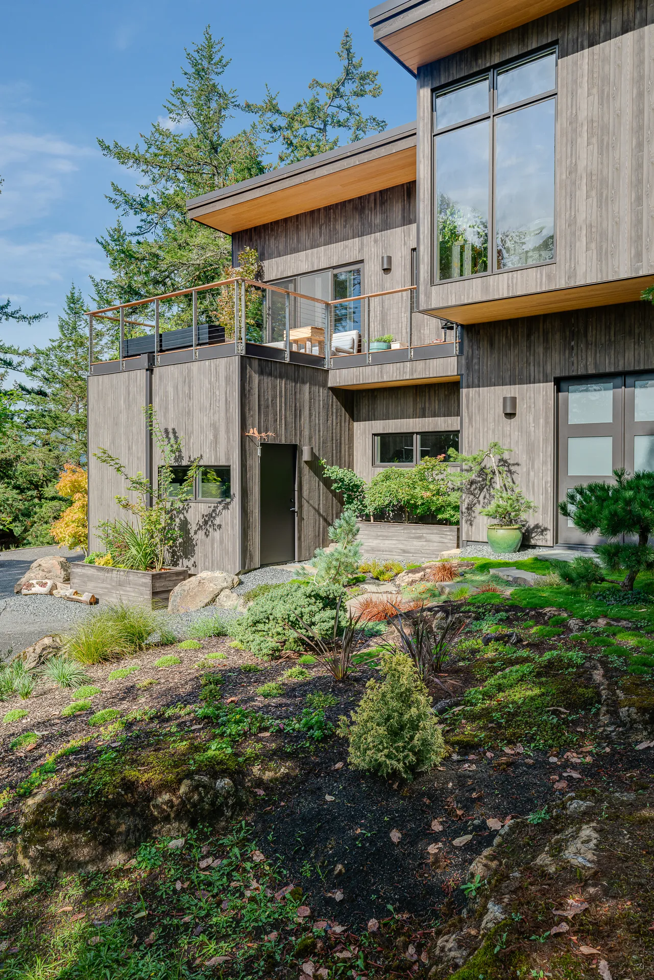 Modern two-story wooden house with large windows, balcony, and landscaped garden with various shrubs and small trees.