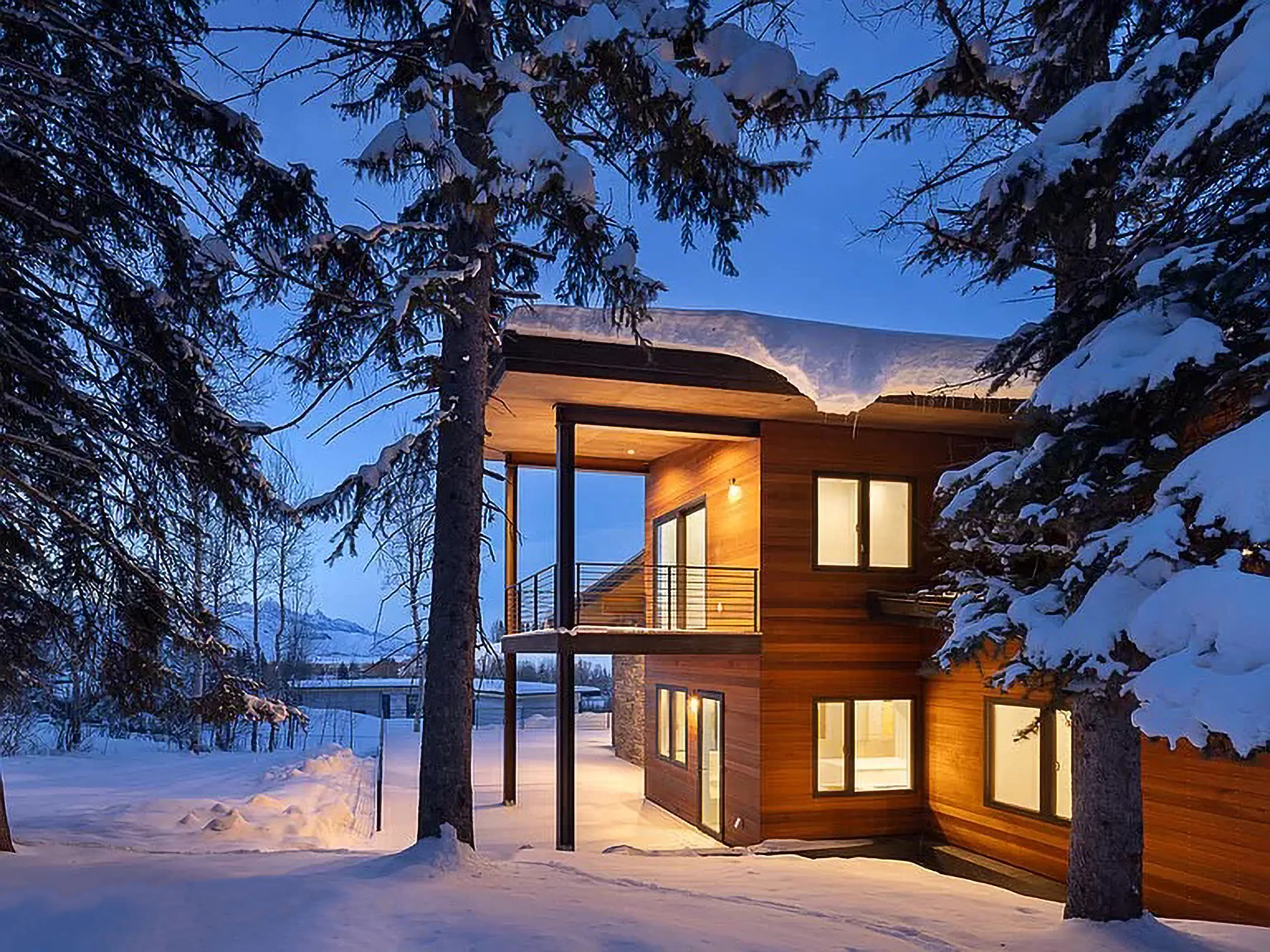 Modern two-story wooden house illuminated at dusk, surrounded by snow-covered trees and snow on the roof.