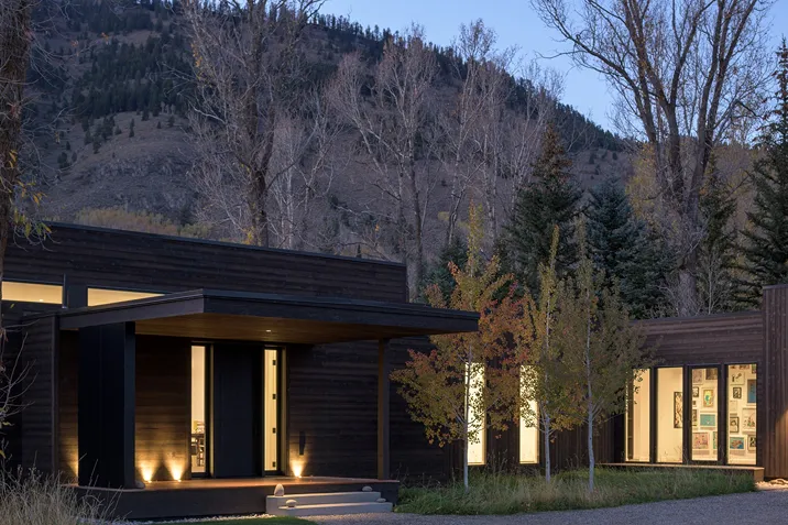 Modern dark wooden house with illuminated windows at dusk, surrounded by trees and a mountain in the background.