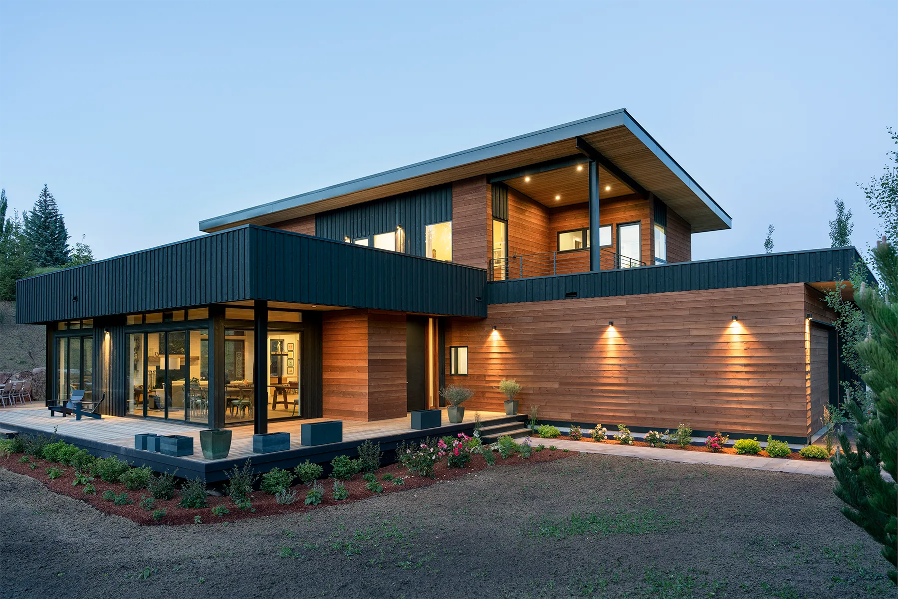 Modern two-story house with wooden and black panel exterior, large windows, and outdoor patio with plants and lighting at dusk.