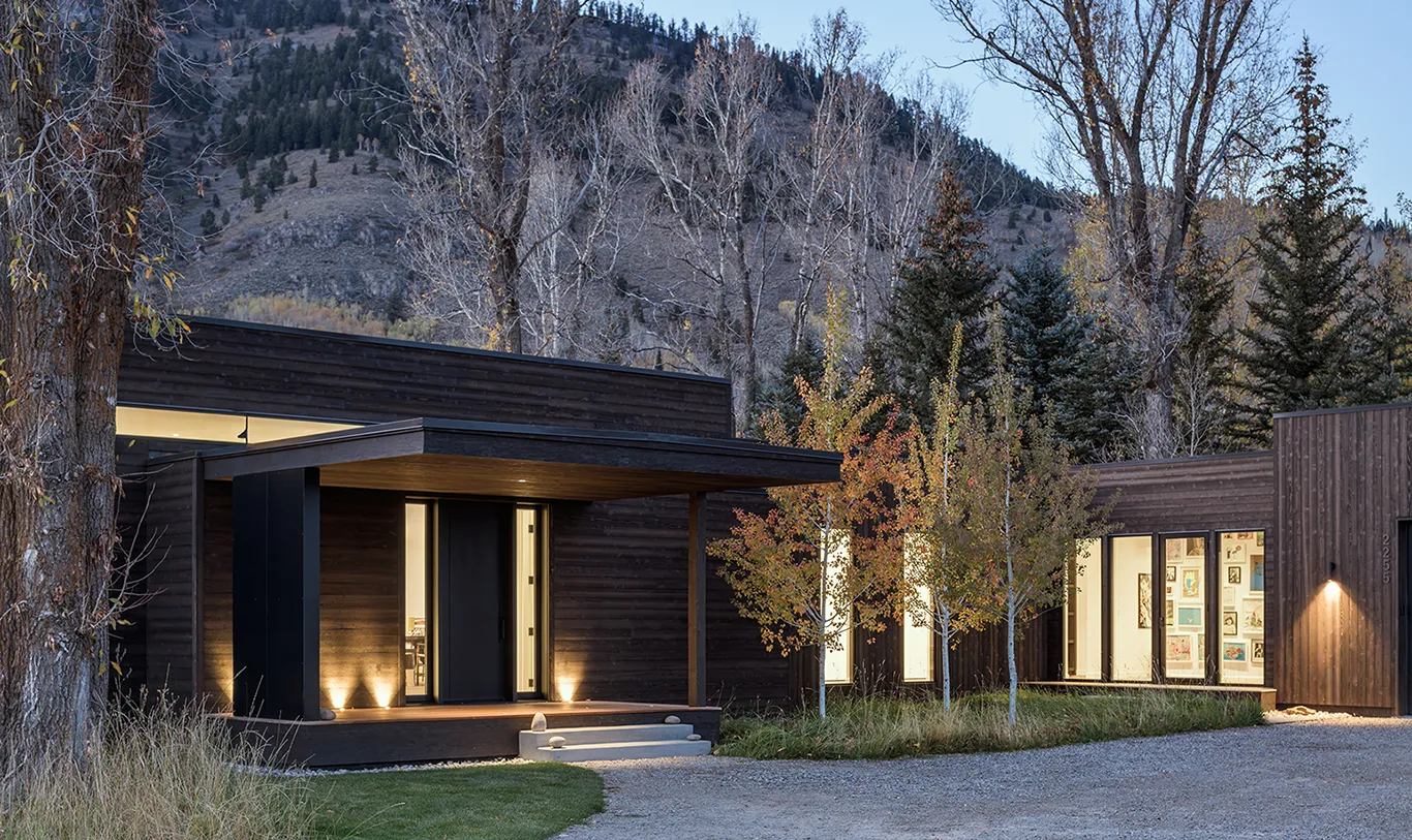 Modern two-story house with wooden and black panel exterior, large windows, and outdoor patio with plants and lighting at dusk.